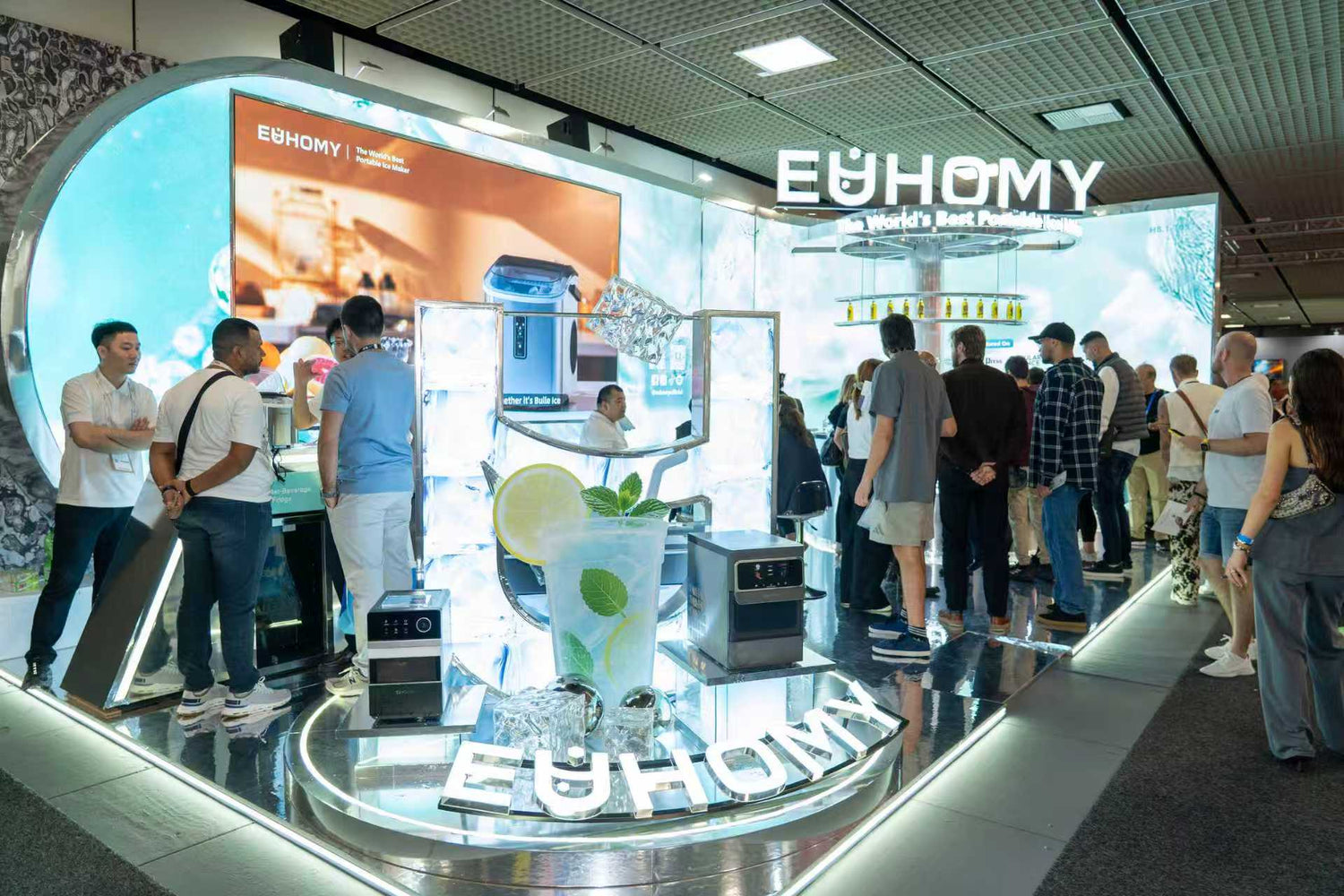 A wide shot of the bustling and futuristic Euhomy booth at CES, with attendees gathered around the illuminated smart ice maker displays.